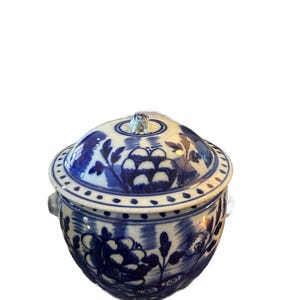 May include: A blue and white ceramic lidded jar with floral designs. The jar has a rounded shape with a lid and small handles. The blue floral pattern is hand-painted on a white background. The lid has a small knob.