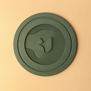 Peut inclure: Un dessous de verre circulaire vert olive présentant un motif abstrait en relief au centre. La surface possède une finition texturée subtile, caractéristique de l'impression 3D. Le motif inclut une forme organique centrale avec deux petits éléments en forme de feuille.