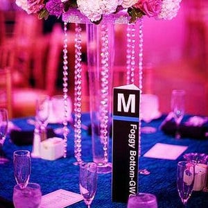 May include: A centerpiece with pink and white flowers, crystal beads, and a clear glass vase. The table is set with blue linens and glasses. A black sign with white text reads "M Foggy Bottom-GW".
