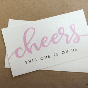 May include: White card with pink cursive text that says "cheers" and black text that says "this one is on us".