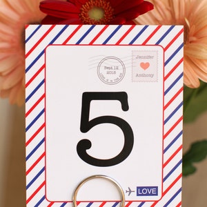 May include: Red, white, and blue striped card with a white background. The card has a large black number 5 on it. The card also has a small stamp with the text "Sept 12 2016 Love is in the air" and another stamp with the text "Jennifer Anthony" with a pink heart. The card has a small airplane icon and the word "LOVE" printed on it.