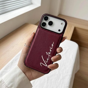 May include: A burgundy phone case with the name "Victoria" in white script. The case has a black border and a camera cutout. The phone case is held by a person.