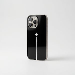 Puede incluir: Funda de teléfono negra con un gráfico de avión blanco y una estela de vapor. La funda tiene un acabado brillante y un anillo de cámara plateado. El diseño es minimalista, ideal para viajeros.