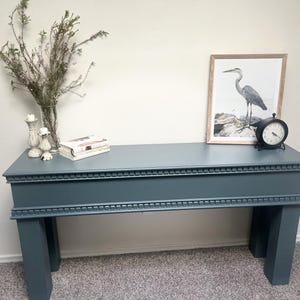 Può includere: Una consolle color teal con finiture decorative, che espone una stampa incorniciata di un airone e un orologio nero. Il tavolo mostra anche una pila di libri, candele e un vaso con fiori secchi. Il tavolo è sostenuto da quattro robuste gambe.