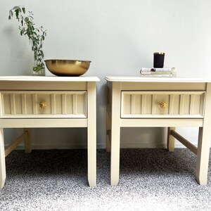 May include: Two cream-colored nightstands with a single drawer each. The drawers have a wood-grain pattern and gold-colored knobs. The nightstands are on a light gray carpet.