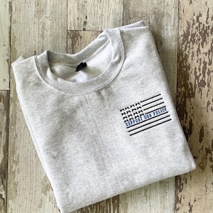 Peut inclure: Un sweat-shirt gris clair avec un col rond. Le motif présente un drapeau noir et blanc avec les mots "SUPPORT OUR POLICE" en bleu. Le sweat-shirt est plié et repose sur une surface en bois.