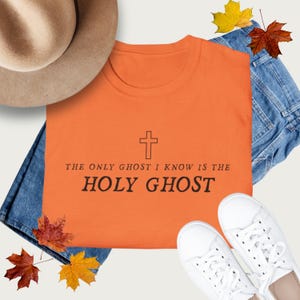 May include: Orange t-shirt with the text "THE ONLY GHOST I KNOW IS THE HOLY GHOST" and a cross symbol. The shirt is laid on top of blue jeans, with a tan hat and white sneakers. Autumn leaves are scattered around the shirt.