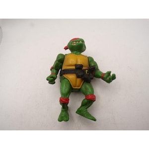 May include: A green action figure of a Teenage Mutant Ninja Turtle, wearing a yellow shell, red head and knee bands, and a black belt with a weapon holster. The figure is posed with arms outstretched.