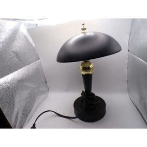 May include: A black desk lamp with a rounded shade and a gold-colored accent. The lamp has a black base and stem, and a black power cord. The lamp is designed in a retro style.