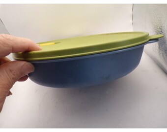 May include: A blue plastic bowl with a green lid. The lid has a small yellow tab. The bowl is held by a hand. The bowl is likely for food storage.