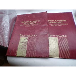 May include: Two maroon New Holland operator's manuals for self-propelled combine models 975 and 985. Each manual features gold text and a gold rectangular graphic at the bottom. The covers show signs of wear and age with visible surface scuffs.