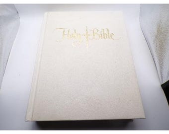 May include: A cream-colored book with the words "Holy Bible" in gold script. The book has a textured cover and a simple, elegant design. The title is centered on the cover.