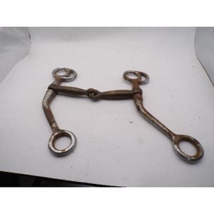 May include: A metal horse bit featuring a jointed mouthpiece with a dark, textured finish and two long, curved shanks. The bit includes four circular rings, with two at the top and two at the bottom, showing signs of oxidation and surface wear.