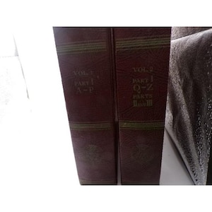 May include: Two dark red, textured hardcover books standing upright. The left spine reads VOL. 1 PART 1 A-P. The right spine reads VOL. 2 PART 1 Q-Z PARTS II AND III. Both spines feature horizontal gold-toned lines and decorative circular emblems.