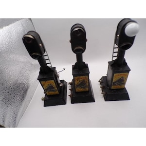 以下が含まれることがあります： 3つのヴィンテージの黒い金属製模型列車信号灯。各信号は長方形のベース、背の高い黒いポスト、白または黒のレンズを備えた信号ヘッドを備えています。ベースには、列車の画像と「AUTOMATIC」という単語が記載された黄色のラベルが付いています。