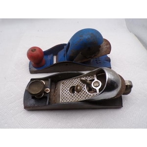 May include: Two hand planes for woodworking. The top plane is blue with a red wooden knob and a metal blade. The bottom plane is silver and black with a textured metal surface, a circular adjustment knob, and a metallic finish on the body.