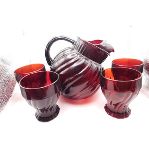 May include: A vintage ruby red glass pitcher and four matching glasses. The pitcher has a large handle and a spiral design. The glasses have a similar spiral design and a footed base. The glassware set is perfect for serving beverages.