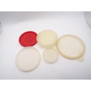 May include: A collection of five plastic lids in various sizes and colors. The lids are white, off-white, and red. The lids are circular and appear to be for food storage containers. The lids are arranged on a white surface.