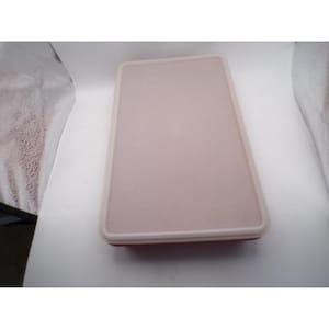 Pode incluir: Um recipiente retangular de plástico rosa claro com uma borda branca. O recipiente tem uma tampa plana e é provavelmente usado para armazenar alimentos. O design é simples e funcional.