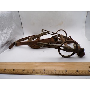May include: A brown leather horse bit with metal cheekpieces and a mouthpiece, positioned behind a wooden ruler that shows the item is approximately 9 inches in length. The metal components have a weathered, dark finish with visible slots.