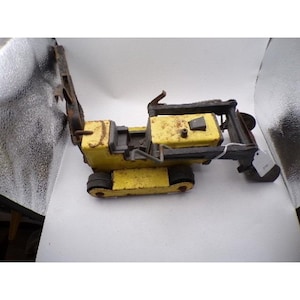 May include: A vintage toy construction vehicle, painted yellow with black accents and rust. The toy features a detailed design of a bulldozer or excavator, with a movable arm and tracks. The toy is likely made of metal and shows signs of wear.