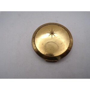 May include: A vintage, round, gold-toned compact with a starburst design on the lid. The compact has a slightly worn appearance, suggesting age and use. The clasp is visible at the bottom. The overall aesthetic is classic and elegant.
