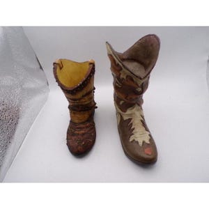 May include: Two miniature cowboy boots. One boot is brown with a yellow interior and studded trim. The other boot is brown and beige with decorative stitching. Both boots have a distressed, vintage appearance.