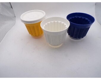 May include: Three plastic coffee filter holders in yellow, white, and blue. The yellow holder has a white lid. The white and blue holders have a ridged design. The holders are sitting on a white surface.