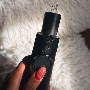 Puede incluir: Un dispositivo electrónico portátil de color negro que presenta una sección superior cilíndrica con varias agujas metálicas finas que sobresalen hacia arriba. El cuerpo incluye un panel de control circular con un botón de encendido y una luz verde.