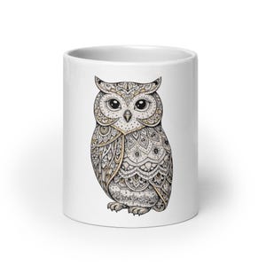 Puede incluir: Taza de cerámica blanca con una ilustración detallada de un búho. El diseño del búho presenta intrincados patrones en tonos beige, dorado y negro. La taza tiene un acabado brillante y una forma cilíndrica simple.