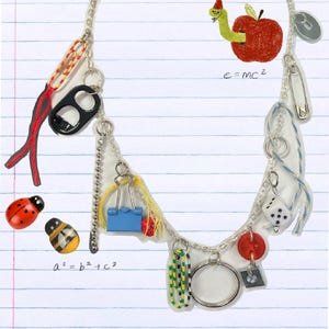 Può includere: Una collana a catena d'argento con vari ciondoli, tra cui un bottone rosso, una graffetta blu, uno spago rosso e giallo, un anello d'argento, un dado, una spilla di sicurezza argentata, una mela rossa con un verme verde, un apribottiglie nero e una coccinella. La collana è su uno sfondo a righe bianche e blu con le equazioni "a^2 = b^2 + c^2" e "e=mc^2" scritte in nero.