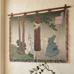 Könnte beinhalten: Ein Wandteppich, der eine japanische Szene mit einer Figur in der Nähe eines Baumes und einer Steinstatue darstellt. Das Kunstwerk ist von einer dunklen Holzstange gerahmt und hat einen Fransenrand. Der Wandteppich hängt an einer beigefarbenen Wand über einem Holzschrank.
