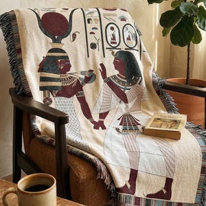 Könnte beinhalten: Beige Decke mit ägyptischem Design, drapiert über einem Holzstuhl. Die Decke zeigt Illustrationen von Figuren, Hieroglyphen und einen Fransenrand. Ein gerahmtes Bild einer Sonne befindet sich an der Wand.