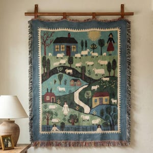 May include: A tapestry depicting a pastoral scene with houses, sheep, and figures, framed by a blue border and fringe. The artwork is mounted on a wooden rod, adding a rustic touch to the wall decor. The color palette includes blues, greens, and earth tones.