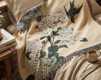 May include: A beige throw blanket with a floral and bird design. The blanket features a fringe trim and is draped over a bed. The floral design is white and green, and the bird is black. A mug of coffee sits on the bed.