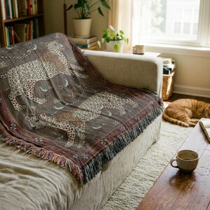 May include: A decorative throw blanket with a leopard print design in shades of brown, tan, and cream, draped over a beige sofa. The blanket has a fringed edge and is placed in a living room setting.