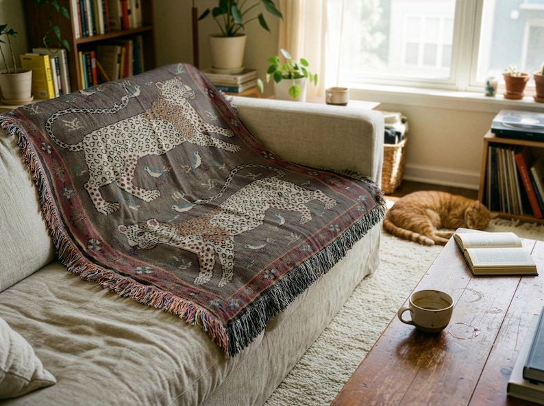 May include: A decorative throw blanket with a leopard print design in shades of brown, tan, and cream, draped over a beige sofa. The blanket has a fringed edge and is placed in a living room setting.