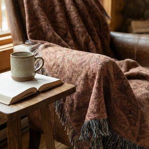 May include: A brown patterned throw blanket with fringe detail draped over a leather armchair. A small wooden side table holds an open book and a ceramic mug. The blanket's pattern has a floral design.