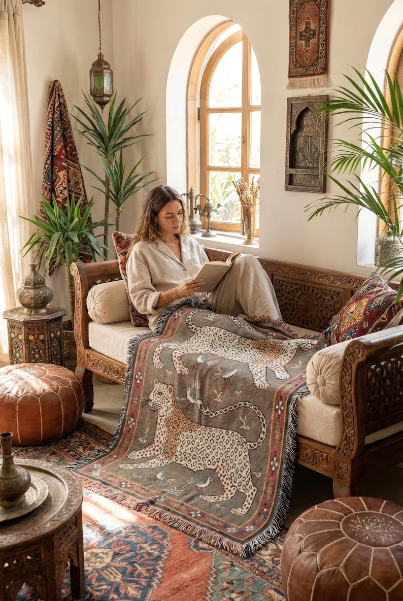May include: A woman reading on a wooden sofa, draped with a gray throw blanket featuring a leopard design. The room has an ornate rug, decorative pillows, and a leather ottoman. The decor has an earthy, bohemian style.