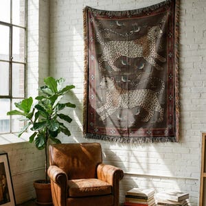 May include: A woven tapestry with a brown background features two running leopards with spotted patterns. The tapestry has a decorative border and fringe. The tapestry is hanging on a white brick wall.