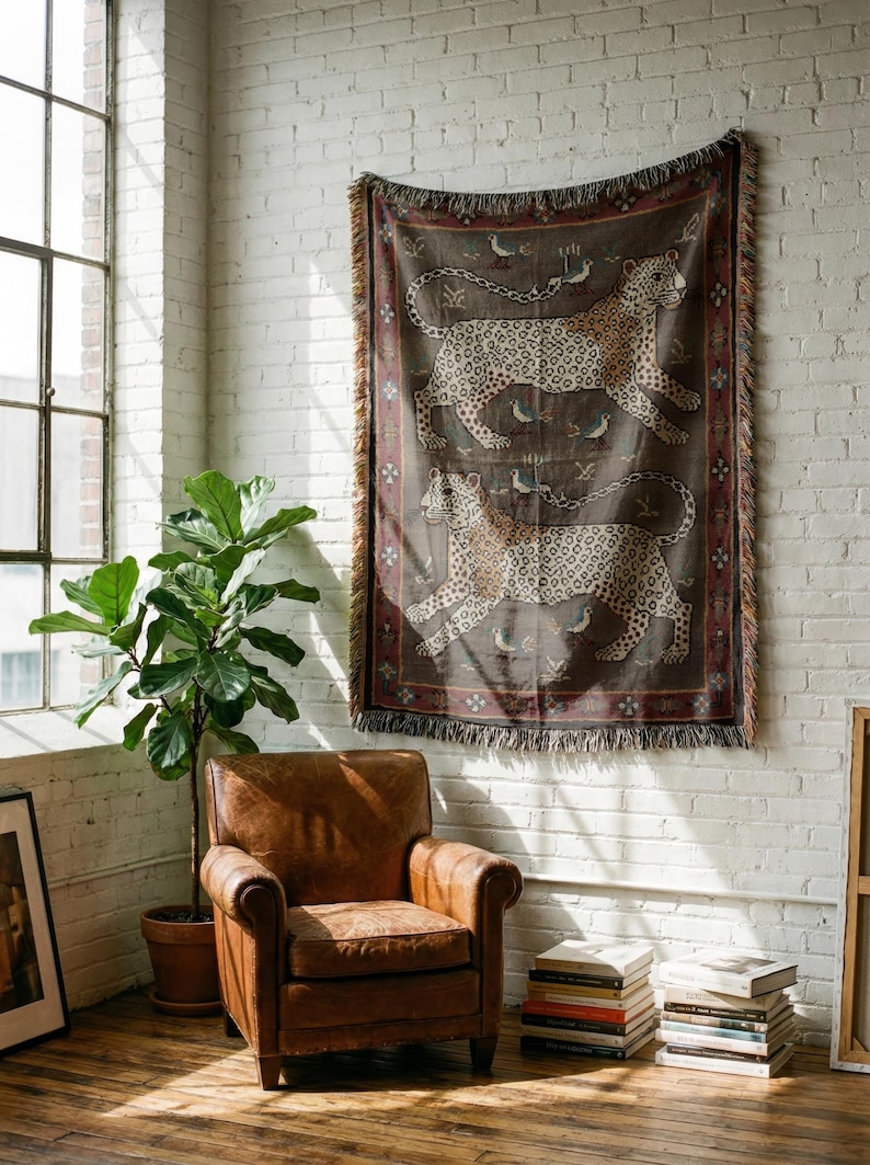 May include: A woven tapestry with a brown background features two running leopards with spotted patterns. The tapestry has a decorative border and fringe. The tapestry is hanging on a white brick wall.