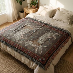May include: A woven throw blanket featuring a repeating pattern of leopards in a neutral color palette. The blanket has a dark gray background with a red border and fringe trim. The throw is displayed on a bed with neutral-colored bedding.