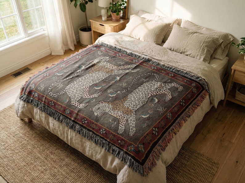 May include: A woven throw blanket featuring a repeating pattern of leopards in a neutral color palette. The blanket has a dark gray background with a red border and fringe trim. The throw is displayed on a bed with neutral-colored bedding.