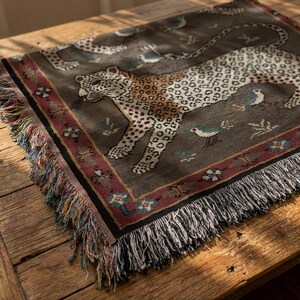 May include: A rectangular throw blanket with a detailed leopard design. The blanket features a dark gray background with a leopard, birds, and floral accents. The border is a burgundy color with a fringe edge.