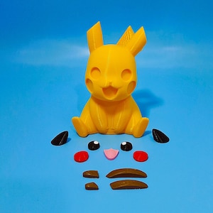 Könnte beinhalten: Eine gelbe, 3D-gedruckte Pikachu-Figur mit separaten schwarzen, roten, rosa und braunen Teilen für Gesicht und Ohren, vor blauem Hintergrund. Die Figur sitzt in einer Position mit lächelndem Gesichtsausdruck.