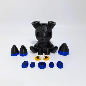 May include: A black 3D-printed creature figurine with separate parts. The figurine has a rounded body, two ears, and is sitting. The parts include blue and black cones, yellow eyes, and blue and black circular pieces.