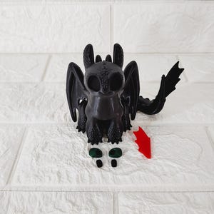 Może przedstawiać: Czarna figurka smoka z dużymi, okrągłymi oczami, skrzydłami i długim ogonem. Figurka stoi na białej powierzchni. Zawiera zielone oczy i czerwoną strzałkę.