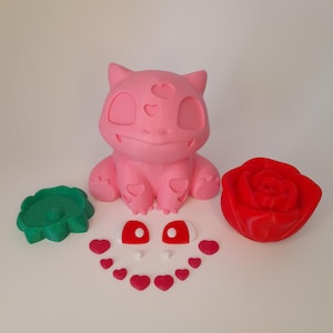 May include: A pink Bulbasaur figure with heart-shaped cutouts, a green circular piece, a red rose, and separate red and white eye pieces, along with small red hearts. The items are arranged on a white surface.