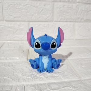 Puede incluir: Una figura azul de Stitch con orejas grandes, ojos negros y vientre azul claro. La figura está sentada con los brazos y las piernas extendidos. Las orejas son rosas por dentro. El fondo es una pared de ladrillos blancos.