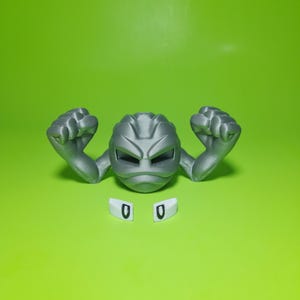 May include: A gray, 3D-printed Geodude figure with raised fists and detached white eye pieces, set against a bright green background. The figure has a detailed, textured surface.